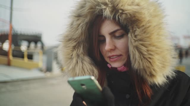 Close-up-shot-of-beautiful-woman-using-smart-phone-technology-app-walking-on-the-street-in-a-warm-jacket-with-a-fluffy-hood