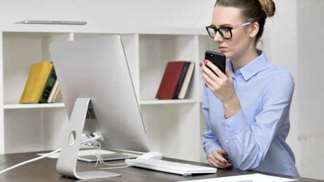 Chica-casual-joven-usando-el-teléfono-inteligente-mientras-trabaja-en-el-ordenador