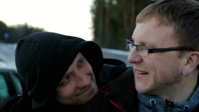 Two-LGBT-guys,-bald-with-a-beard-and-wearing-glasses-and-a-hat-hugging-and-chatting-on-the-side-of-the-road-near-the-car