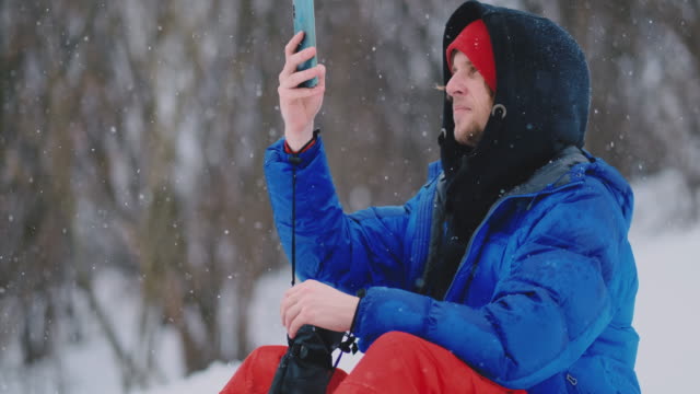 Male-snowboarder-sitting-on-the-snow-taking-photos-on-the-phone-beautiful-scenery-of-the-resort-for-social-networks