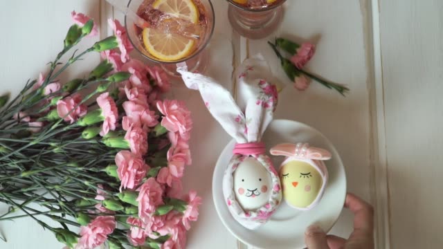 Female-hand-puts-plate-with-Easter-egg-decorated-for-Easter-bunny-and-Easter-chick,-against-background-of-pink-carnations-and-lemonade