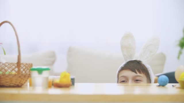 Young-adorable-boy-is-hiding-under-the-table-full-of-Easter-decorations-and-is-playing-with-Easter-bunny-in-his-hand.-Bunny-theatre.-Discussion.-Conversation:-cheerful-boy-and-a-soft-bunny.