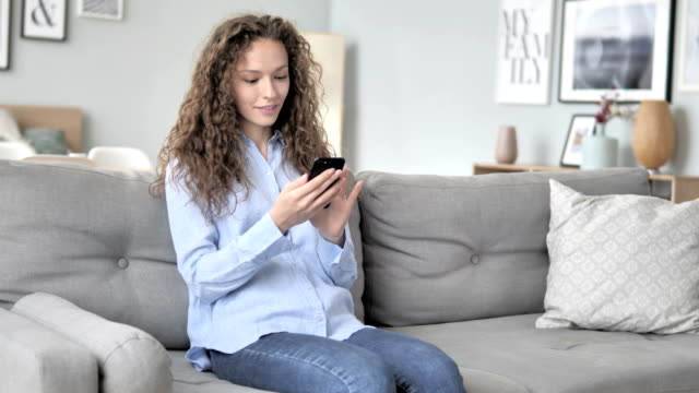 Young-Curly-Hair-Woman-Using-Smartphone-while-Relaxing-on-Sofa