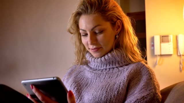 Closeup-shoot-of-adult-caucasian-blonde-female-browsing-on-the-tablet-and-cheerfully-smiling-looking-at-camera-indoors-in-a-cozy-apartment