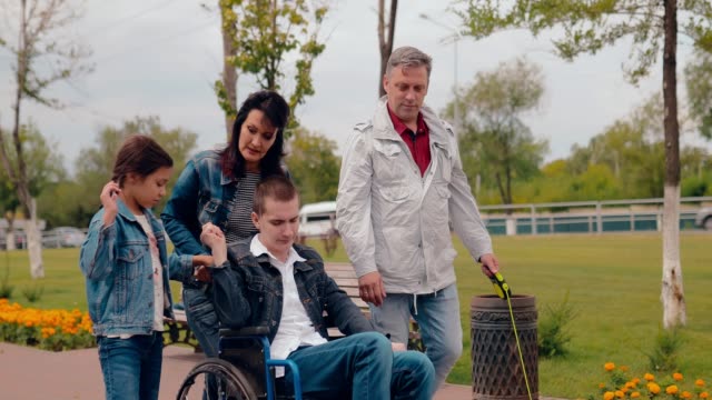 Schwester-und-Mutter-nehmen-Bruder-in-den-Rollstuhl.-Glückliche-Familie-mit-behinderten-Teenager-zusammen-zu-Fuß.