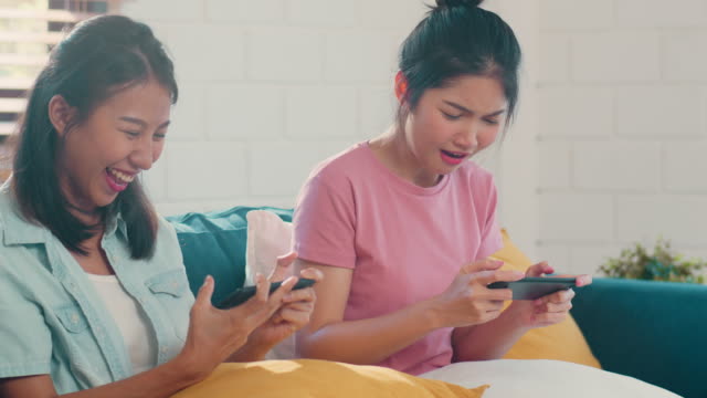 Junge-asiatische-lesbische-Paar-mit-Handy-spielen-Spiele-im-Internet-zusammen,-während-liegen-Sofa-im-Wohnzimmer.