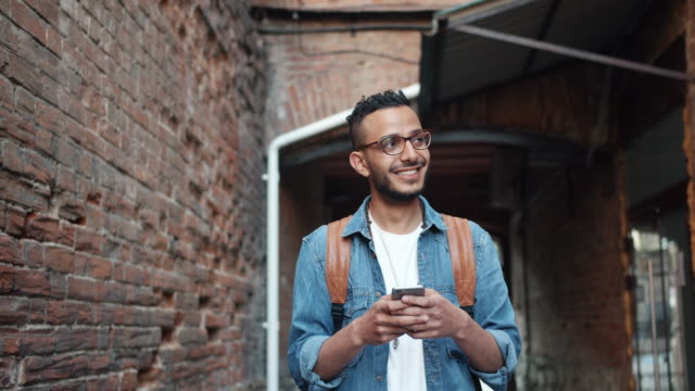 Guapo-árabe-barbudo-disfrutando-de-la-aplicación-de-teléfono-inteligente-moderno-sonriendo-al-aire-libre