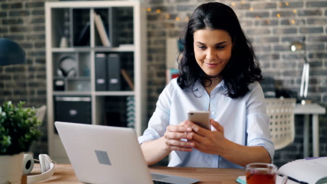 Pretty-girl-touching-smartphone-screen-and-smiling-sitting-at-desk-in-office