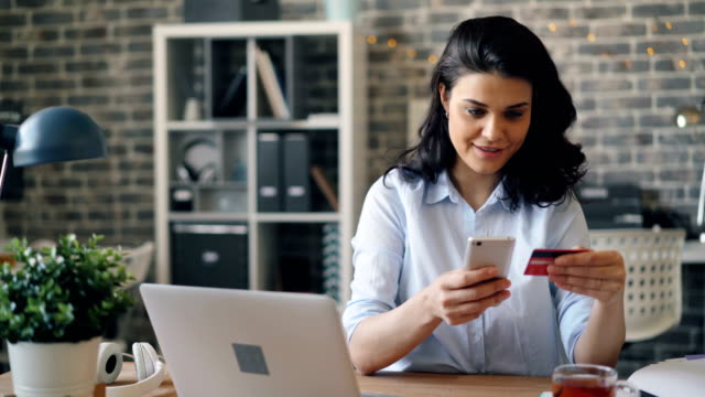 Young-woman-paying-online-with-credit-card-using-smartphone-in-workplace