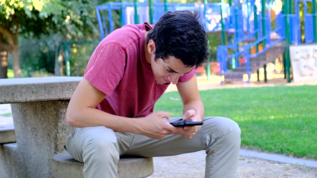 Junger-Mann-mit-seinem-Smartphone-sitzt-in-einer-Parkbank