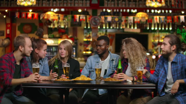 Multiethnische-Gruppe-junger-Männer-und-Frauen,-die-an-einer-Bar-Bier-trinken-und-eine-lustige-Diskussion-über-die-Universität-führen
