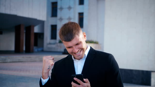 Businessman-celebrating-success.-Man-received-a-message-with-good-news.-Successful-Businessman-showing-Yes-gesture,-extremely-happy-about-good-news-from-smartphone.-Office-building-background.