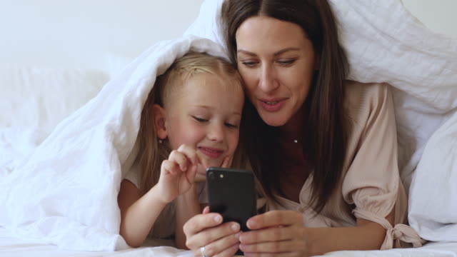 Glückliche-Mutter-und-Kind-Tochter-liegen-auf-dem-Bett-mit-Telefon