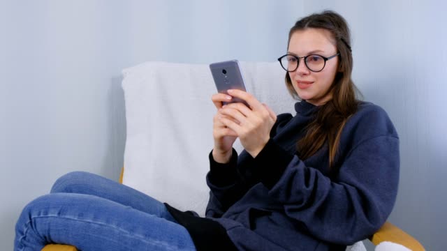 Young-brunette-woman-in-glasses-types-a-message-on-a-mobile-phone-sitting-in-armchair.