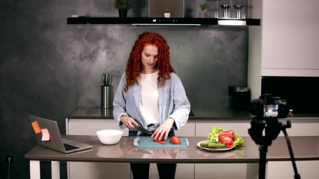 Young-woman-in-casual-clothes-leads-a-culinary-blog-and-records-video-in-grey-kitchen.-Video-shooting-of-the-blog---red-headed-cutting-tomatos-on-a-dashboard.-Modern-marketing,-social-networks