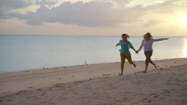 Zwei-junge-glückliche-Frauen-laufen-am-Strand-bei-Sonnenuntergang