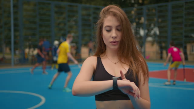 Close-up-shooting.-Portrait-of-a-sports-girl.-The-girl-is-looking-at-the-training-information-on-her-smartwatch.-People-are-playing-floorball-in-the-background.-4K