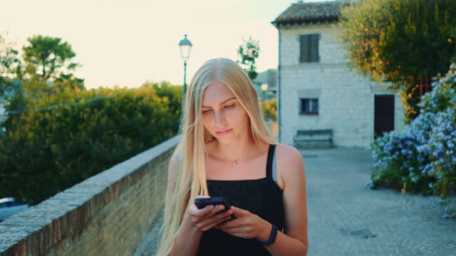 Charming-young-woman-using-smartphone-while-walking-outside-in-summer