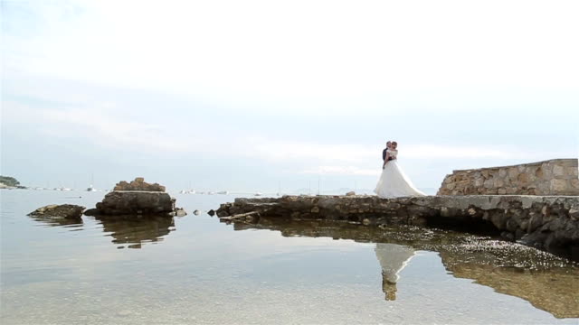 Man-and-woman-in-formal-clothes-kiss-at-old-pier-by-sea.-Groom-walks-to-bride-in-wedding-dress-embraces-her-standing-at-ancient-castle-ruins-dock-near-lake-water-reflection---yacht-boats-background