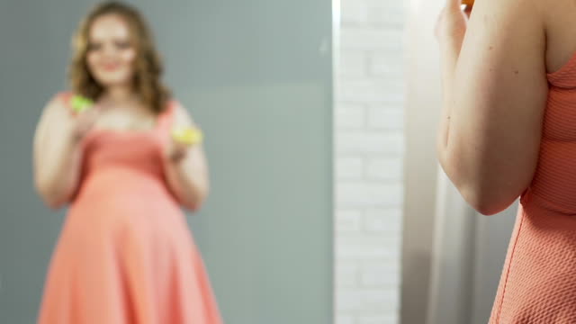 Mujer-con-sobrepeso-en-el-vestido-de-comer-donuts-admirando-su-reflejo-en-el-espejo