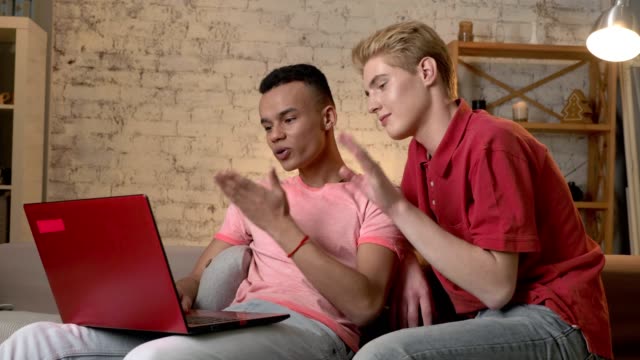 Two-multinational-homosexual-friends-sit-on-the-couch,-use-a-laptop,-show-a-gesture-of-goodbye,-air-kiss-in-a-video-chat.-Home-cosiness,-family,-internet-concept.-60-fps