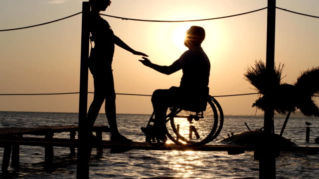 romantisches-Date-weiblich-mit-männlichen-ungültig-auf-Rollstuhl-am-Steg-in-der-Nähe-von-Meer-gegen-Sommer-Sonnenuntergang