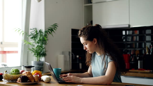 Young-concentrated-cheerful-woman-working-in-kitchen-typing-laptop-computer-during-breakfast-in-the-morning