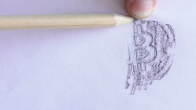 Young-male-professional-is-drawing-bitcoin-image-on-white-paper-using-pencil,-on-table-in-trader-company,-person-depicts-symbol-of-peering-payment-system-on-sheet-on-desk-in-room-with-daylight