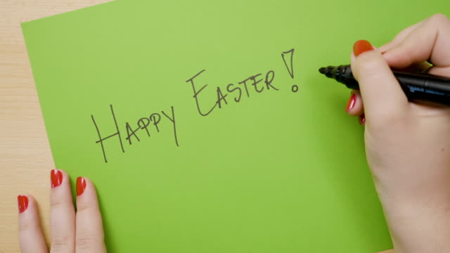 Woman-hands-with-red-manicure-writing-happy-easter-in-capital-letters-on-green-paper-with-a-black-marker