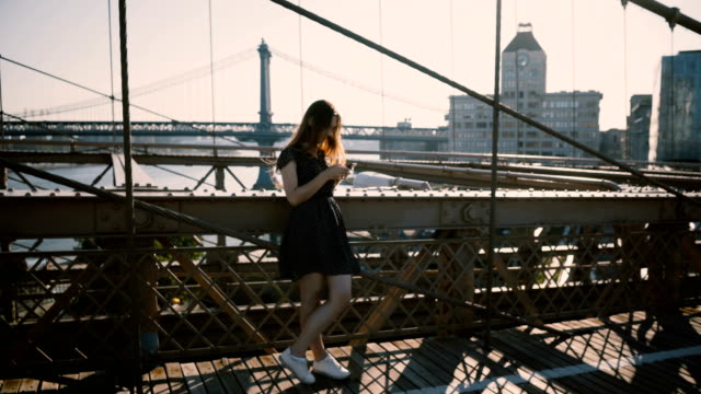 Hermosos-soportes-de-mujer-caucásica-con-aplicación-de-mensajería-de-smartphone-en-el-puente-de-Brooklyn,-Nueva-York-increíble-panorama-horizonte-4K