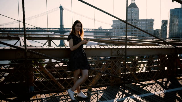 Young-pretty-European-girl-looking-around,-using-smartphone-app-at-Brooklyn-Bridge,-New-York-amazing-riverside-view-4K