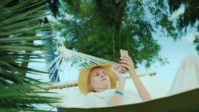 Young-woman-resting-in-a-hammock,-using-a-mobile-phone.-Connection-on-vacation-concept