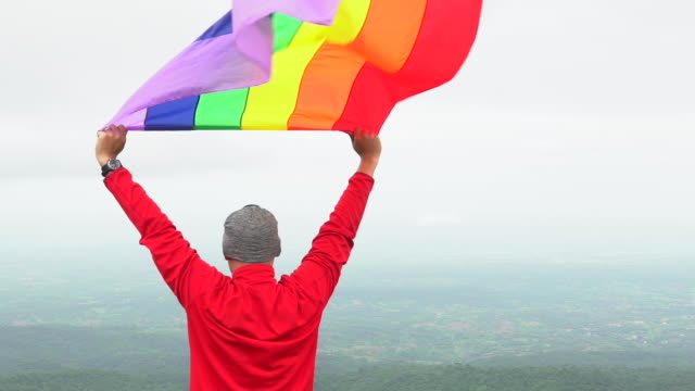 Mann-erhöhen-Regenbogen-Farbe-LGBTI-Fahnenschwingen-in-harten-Wind-auf-Mountain-Top-Sicht