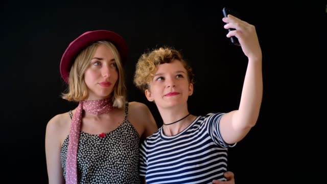 Zwei-charmante-junge-blonde-Frauen,-die-isoliert-umarmt-und-nehmen-Selfie,-stehend-auf-schwarzem-Hintergrund
