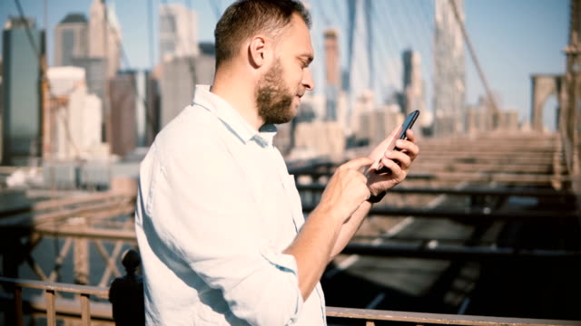 Happy-European-businessman-using-smartphone-mobile-office-app-at-Brooklyn-Bridge,-then-turning-away-from-camera-4K