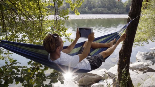 Hombre-joven-con-tableta-digital-en-hamaca,-mintiendo-entre-los-árboles-por-el-río-en-verano