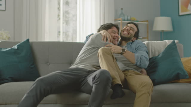 Sweet-Male-Gay-Couple-Full-Around-on-a-Sofa-at-Home.-Boyfriend-Runs-and-Jumps-into-Hands-of-His-Partner.-They-Hug-and-Look-at-Each-Other.-They-are-Happy-and-Laughing.-Room-Has-Modern-Interior.