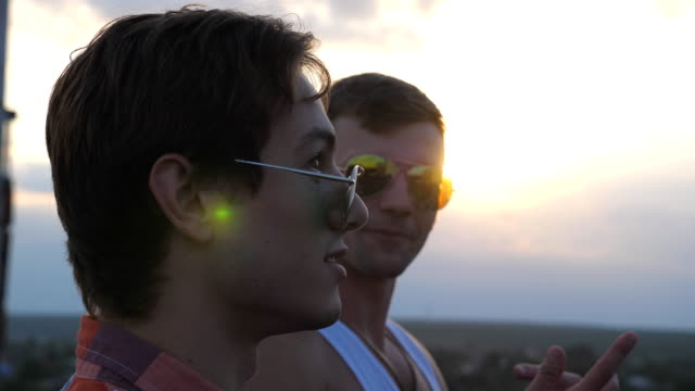 Profile-of-young-male-couple-in-sunglasses-standing-on-the-edge-of-rooftop-and-talking.-Handsome-gay-boys-relaxing-on-roof-of-high-rise-building-and-enjoying-beautiful-cityscape.-Close-up-Slow-motion