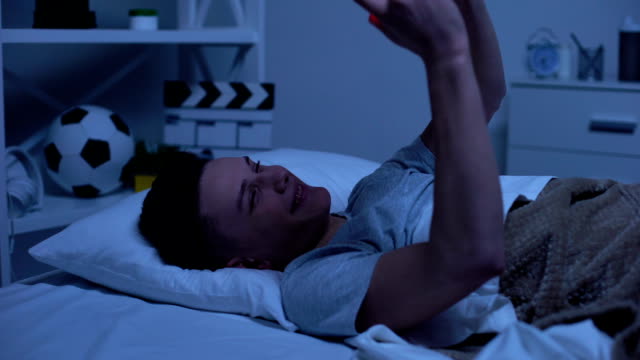 Teen-boy-making-selfie-on-smartphone-putting-it-in-bed-and-falling-asleep,-relax