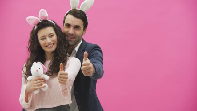 Young-couple-standing-on-a-pink-background.-During-this-show-a-gesture-class-smiling-and-holding-a-soft-toy-in-the-hands-of-a-bunny.-Happy-family-is-preparing-for-Easter,-with-the-ears-of-a-pink-rabbit-on-its-head.
