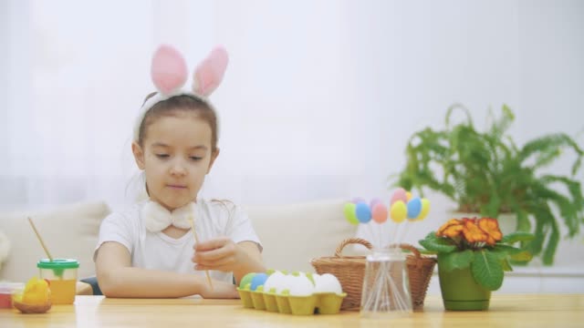 Little-cute-girl-is-having-fun-holding-a-paint-brush-in-her-left-hand.-Girl-with-a-beauty-spots-at-her-face-watching-at-nude-paint-brushes,-sitting-at-the-wooden-table-with-Easter-decorations.-Girl-shows-her-fingers-up.