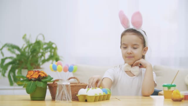 Little-cute-and-adorable-girl-is-smiling-sincerely.-She-takes-an-Easter-egg-from-the-basket-and-looking-at-it,-then-pointing-on-it.-Concept-Easter-holiday.