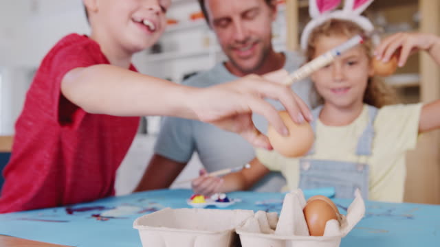 Padre-con-niños-vistiendo-orejas-de-conejo-decorar-huevos-de-Pascua-en-casa-juntos