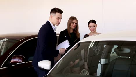 Two-luxurious-young-girls-in-a-car-dealership-talk-to-a-car-salesman.-Car-sale.