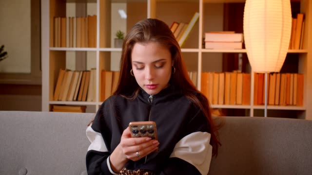 Portrait-of-young-teenage-girl-watches-into-cellphone-sitting-on-sofa-on-bookshelves-background-at-home.