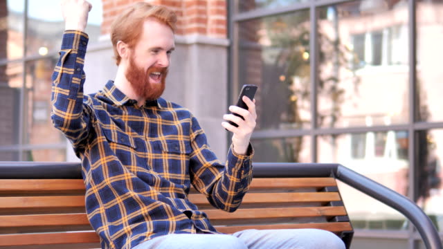 Rotschopf-Bart-junger-Mann-feiert-Sieg-auf-dem-Smartphone,-sitzen-im-Freien-auf-Bank