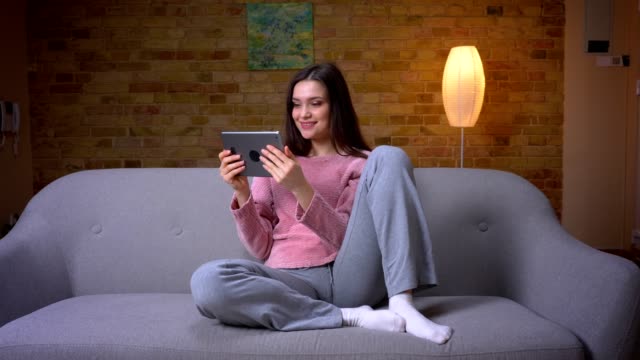 Closeup-shoot-of-young-pretty-brunette-caucasian-female-texting-on-the-tablet-sitting-on-the-couch-in-a-cozy-apartment-indoors
