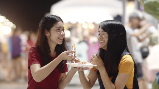 Asian-young-women-friends-travel-in-Bangkok,-Thailand,-beautiful-female-feeling-happy-walking-and-eating-Pad-Thai-at-The-Khao-San-Road.-Women-travel-eat-street-food-concept.-Slow-motion-shot.