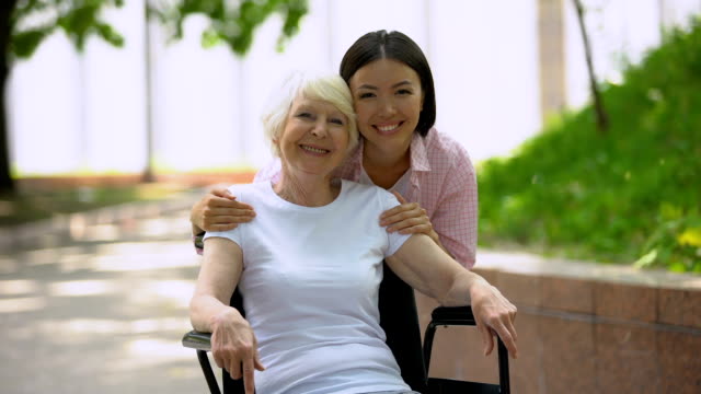 Mujer-joven-abrazando-a-la-abuela-en-silla-de-ruedas-y-mirando-la-cámara-al-aire-libre
