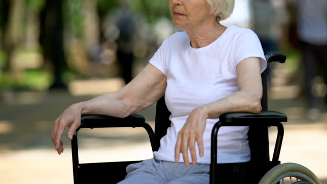 Traurige-Seniorin-im-Rollstuhl-sitzt-im-Park-und-schaut-auf-Menschen,-Behinderung